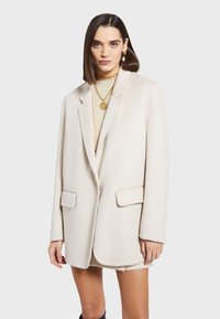 Blazer de color crema de gran tamaño con solapas de muesca, dos bolsillos frontales y una textura suave, usado sobre un suéter ligero y joyas de oro.