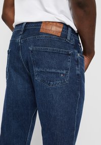 Jeans de mezclilla azul oscuro con un acabado texturizado. Presentan un parche de cuero con el logo en la cinturilla y bolsillos traseros con sutiles detalles de costura.