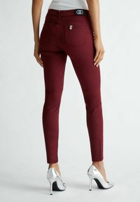 Femme portant un pantalon slim bordeaux et des chaussures à talons hauts argentées, debout les mains détendues le long du corps devant un fond uni.