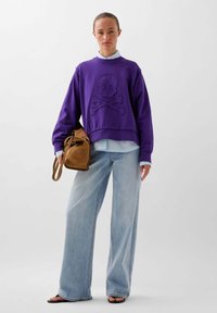 Paarse sweatshirt met een ontwerp van een doodshoofd en gekruiste botten, gedragen over een lichtblauw shirt, gecombineerd met vervaagde wijde pijpen jeans en een bruine schoudertas.