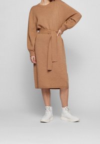 Strickkleid in Hellbraun mit langen Ärmeln, tailliert mit einem Gürtel und in knielanger Länge; kombiniert mit weißen Schnürstiefeletten.