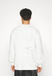 Bílá mikina s texturovaným povrchem, žebrovanými manžetami a spodním lemem, a kulatým výstřihem, zobrazená ze zadní strany.