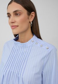 Blusa azzurro chiaro con righe verticali, frontale plissettato, colletto alto e bottoni decorativi a forma di fiore in tonalità argento sulla spalla. Texture del tessuto liscia.