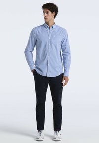 Knoopblouse met blauw-witte verticale strepen, lange mouwen en een borstzak, gecombineerd met donkere broek en witte sneakers.