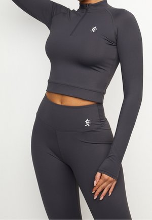 Kobieta nosząca ciemnoszarą dopasowaną bluzkę typu crop top z długim rękawem i zamkiem błyskawicznym oraz dopasowane legginsy z wysokim stanem z małymi białymi logo.