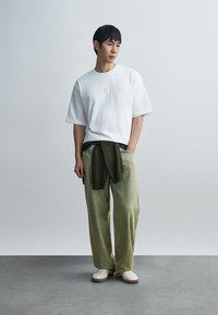 Λευκό πουκάμισο με κοντά μανίκια και λογότυπο στο στήθος, φαρδιά παντελόνια με πράσινες ρίγες και σκούρο πράσινο πουλόβερ δεμένο στη μέση. Κρεμ αθλητικά παπούτσια.