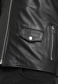 Giacca di pelle nera con zip argentata, tasca con pattina e bottone a pressione, e tasca laterale con zip, indossata sopra jeans neri.