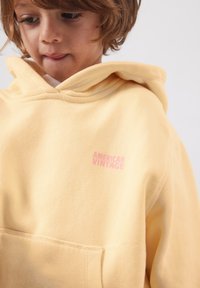 Jeune enfant aux cheveux châtain clair portant un sweat à capuche jaune pâle avec une poche avant et le logo "American Vintage" sur la poitrine.