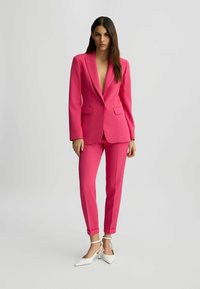 Ensemble de costume ajusté fuchsia comprenant un blazer à col en V profond, deux poches avant et un pantalon slim ; assorti de chaussures blanches à talons pointus.