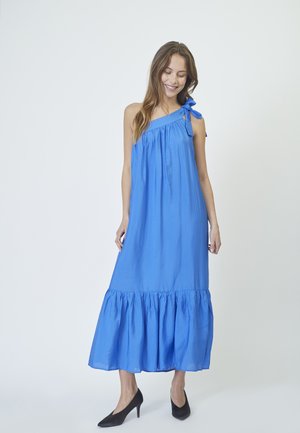 Abito maxi blu senza maniche con corpetto arricciato e orlo a strati. Il cinturino sinistro presenta un dettaglio a fiocco. La modella indossa scarpe con tacco nere.