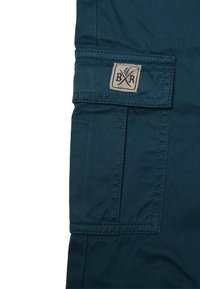 Mörk teal cargobyxfabrik med synliga sömmar, en sidoficka och en beige etikett med korsade verktyg och "BR". Slät textur.