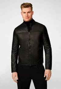 Zwarte leren jas met staande kraag, ritssluiting aan de voorkant en twee zijzakken. Gladde textuur, strakke pasvorm en minimalistische stikseldetails.