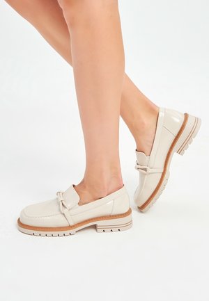 Pieds portant des mocassins couleur crème avec des semelles épaisses marron clair sur fond blanc.