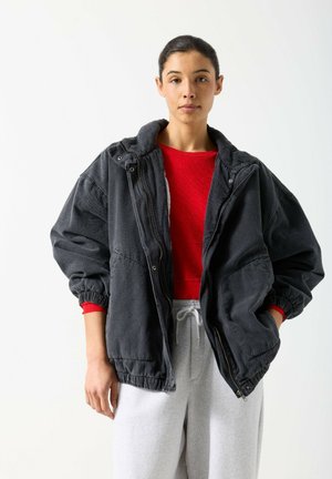 Jeune femme aux cheveux foncés attachés, portant un haut rouge, une veste noire surdimensionnée et un pantalon de survêtement gris clair, debout devant un fond blanc.