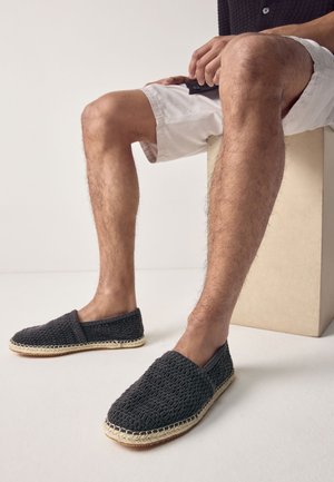 Uomo seduto su un blocco beige, indossa pantaloncini bianchi e mocassini intrecciati neri con suole beige cucite.