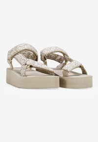 Sandalias de plataforma con una correa de tela de un beige claro que presenta un patrón geométrico, y un tacón grueso sólido de color beige. Superficie texturizada con detalles en dorado.