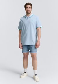 Jasnoniebieska koszulka polo z zielonymi akcentami i krótkimi rękawami, w zestawieniu z dżinsowymi szortami i białymi trampkami. Prosty, casualowy design.