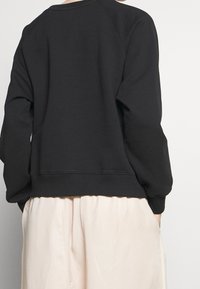 Felpa nera con maniche raglan, colletto e bordo a coste, realizzata in tessuto morbido. Indossata sopra pantaloni chiari e larghi.