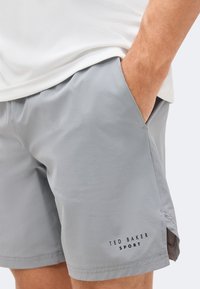 Άνδρας που φοράει ανοιχτό γκρι αθλητικές βερμούδες Ted Baker με το χέρι στην τσέπη και ένα λευκό πουκάμισο μερικώς ορατό.