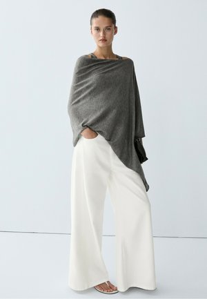 Frau steht und trägt einen grauen asymmetrischen Poncho, weiße Hosen mit weitem Bein und weiße Sandalen, mit einer kleinen schwarzen Tasche in der einen Hand.