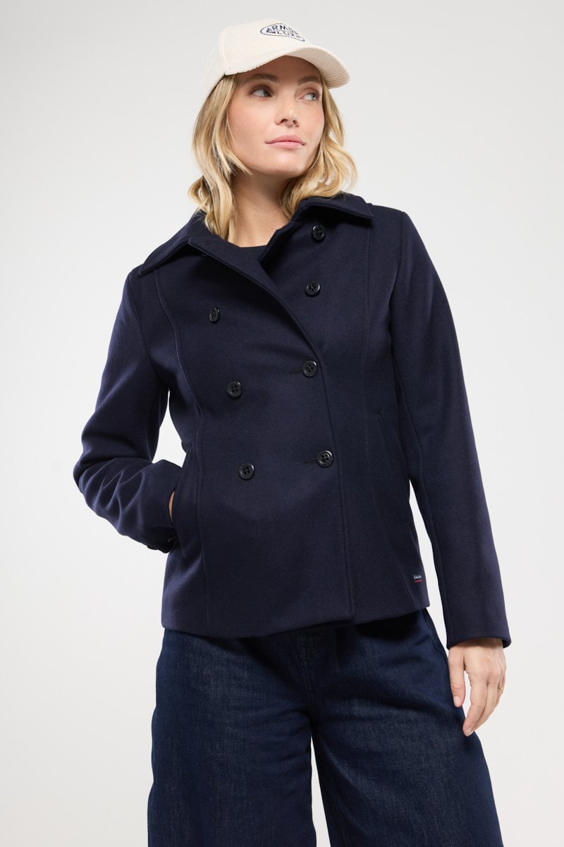 Manteau bleu marine à double boutonnage avec larges revers, fermeture à boutons et deux poches latérales. Associé à un jean large bleu foncé et une casquette de couleur claire.