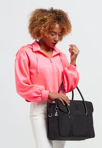 Schwarze Tragetasche mit Klappe, Ledergriffen und einem Schlüsselanhänger-Detail. Kombiniert mit einer hellrosa Satin-Jacke mit Reißverschluss und weißen Hosen.