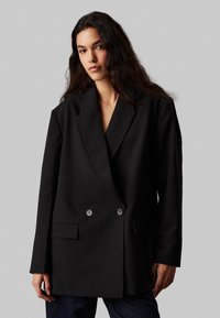 Blazer nero doppiopetto realizzato in un tessuto liscio, con rever a doppio spacco, due tasche frontali e dettagli con bottoni.