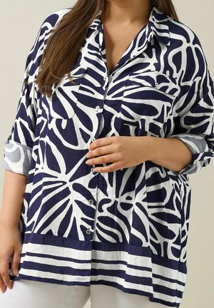 Frau trägt ein locker sitzendes, navyblau-weiß gemustertes Hemd mit aufgekrempelten Ärmeln und weiße Hosen, Hand ruht in der Nähe der Knöpfe.