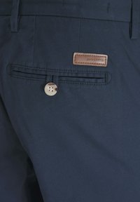 Pantaloni blu navy con una texture liscia, dotati di un'etichetta in pelle sulla tasca posteriore e una chiusura a bottone sulla tasca.