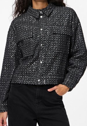 Lähikuva henkilöstä, joka käyttää mustaa paljettitakin, jossa on kaksi rintataskua ja mustat housut, käsi taskussa.