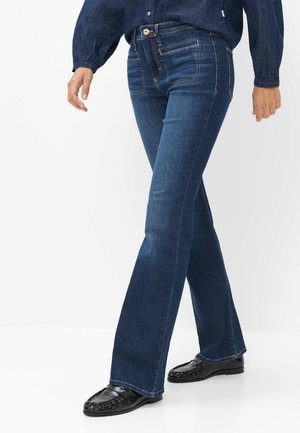 Person iført mørkeblå denimjeans og sorte lædermokkasiner, der står med det ene ben krydset foran det andet.
