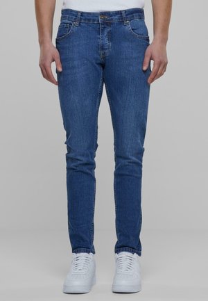 Mann trägt Slim-Fit-Blaue Jeans und weiße Sneakers, steht vor einem schlichten grauen Hintergrund.