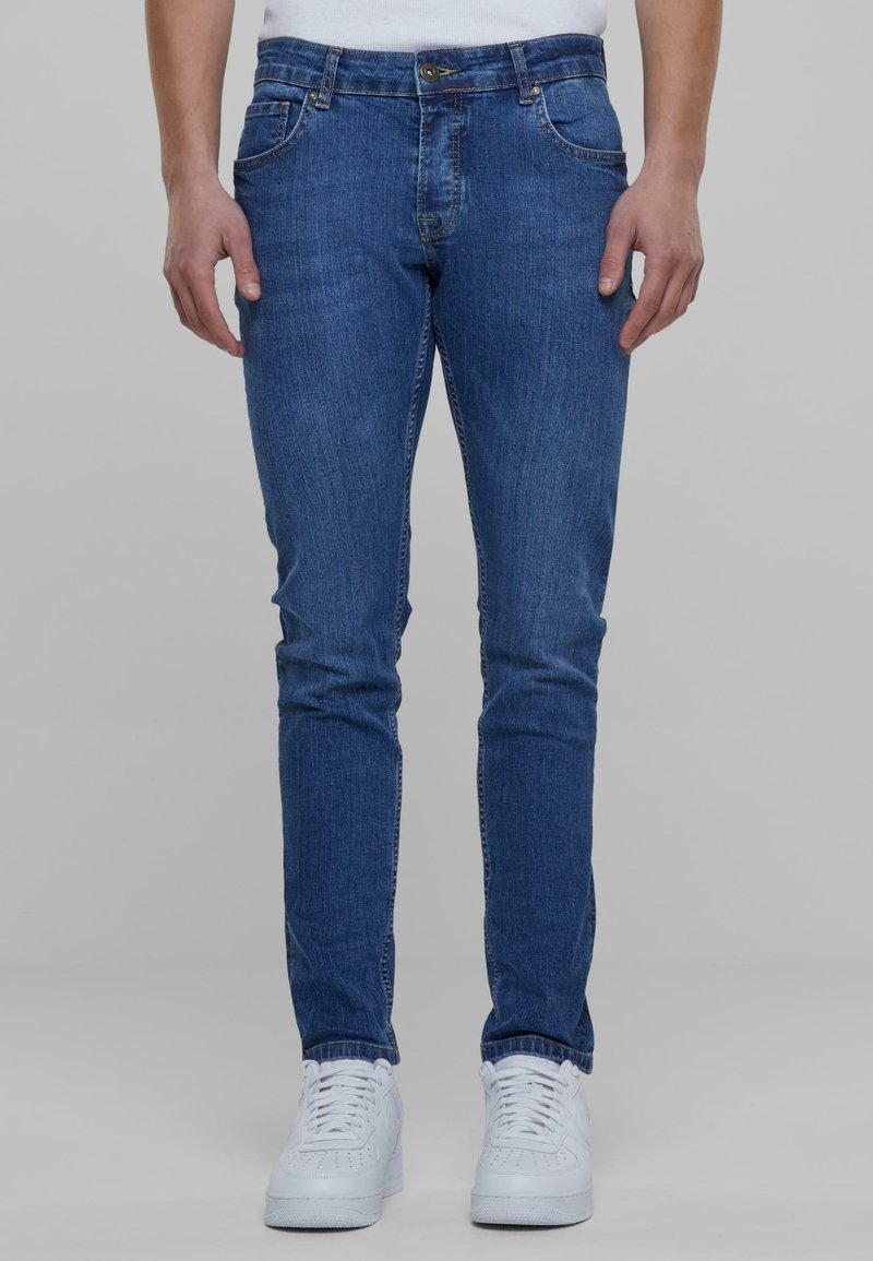 Homme portant un jean bleu slim et des baskets blanches, debout devant un fond gris uni.