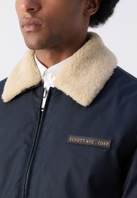 Chaqueta bomber navy con un cuello de borreguito beige, que presenta un cierre de cremallera y una parche de cuero marrón etiquetado "SCHOTT NYC. CORP."