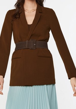 Femme portant un blazer marron avec une large ceinture marron foncé et une jupe plissée bleu clair, avec des colliers dorés superposés visibles.