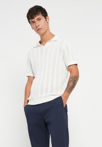 Maglione bianco a maniche corte con colletto, caratterizzato da un motivo tessuto a maglia e orlo a coste; abbinato a pantaloni blu navy.