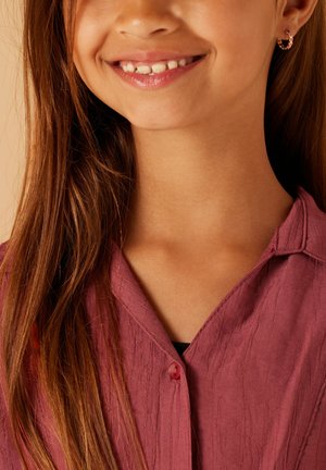 Chemise boutonnée en rose poussiéreux avec une surface texturée. Gros plan sur le col et le sourire, mettant en avant de longs cheveux bruns et des boucles d'oreilles en or.