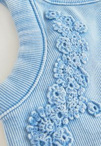 Helles blaues ribbeltes Material mit strukturierten Blumenstickereien auf einer Seite. Glatte Kanten mit einem runden Ausschnitt und sanftem Schimmer.