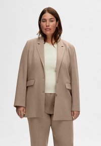 Beige skräddarsydd blazer med en enda knappstängning och två framfickor, bärs över en ljusgrön ribbad topp, matchande beige byxor.