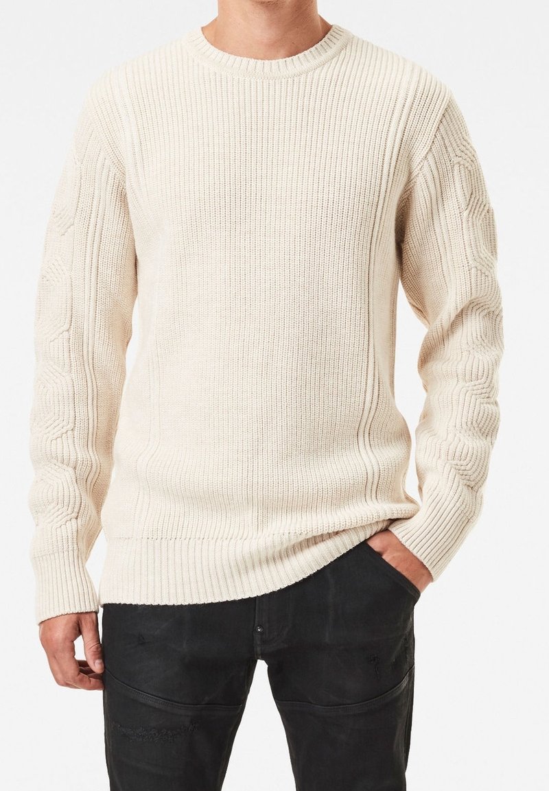 Cream sweater with ribbed texture and cable knit details on sleeves. Crew neck design, long sleeves, paired with dark trousers.