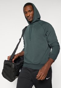 Tmavě zelená mikina s kapucí, stahovací šňůrkou, raglánovými rukávy a logem Nike na hrudi, doplněná černými sportovními kalhotami a černou taškou.