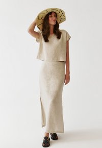 Linen matching two-piece set: cropped top with relaxed sleeves and a long, side-slit skirt. Soft beige color. Worn with black sandals and wide-brimmed hat.