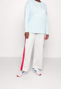 Ljusblå sweatshirt med vitt Adidas-logotyp, ihop med vita byxor med röda och blå sidostrikar. Vita skor med röda detaljer.