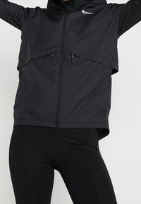 Veste coupe-vent Nike noire avec zip sur le devant, capuche et design en superposition. Présente un logo contrasté sur la poitrine et des poches zippées sur les côtés.
