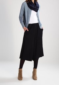 Vaaleansininen avonainen neulecardigan, jossa on pyöreä kaula-aukko, tummansininen huivi, valkoinen toppi, mustat leveät housut ja beige nilkkabootsit. Suuri tekstuuri ja rento tyyli.