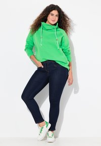 Sudadera verde con cuello tipo cowl, cordones blancos y detalles dorados, combinada con jeans ajustados azul oscuro y zapatillas blancas con acentos verdes.