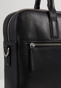 Coin d'une mallette en cuir noir avec une fermeture éclair argentée, une poche avant, des poignées cousues et une attache en métal sur un fond gris clair.