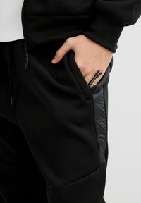 Pantalón jogger negro con tejido texturizado, bolsillos ajustados y detalles brillantes en los laterales. Detalle de cremallera en el bolsillo con cierre de cordón en la cintura.