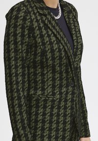 Teksturowana czarno-zielona marynarka w kratę z jednym zapięciem na guzik, klapami z wycięciem oraz detalem z kieszenią z przodu.