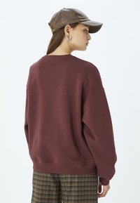 Sudadera oversized burdeos con puños y dobladillo acanalados. Combinada con una gorra de cuero marrón y pantalones de cuadros, mostrando un ajuste relajado.
